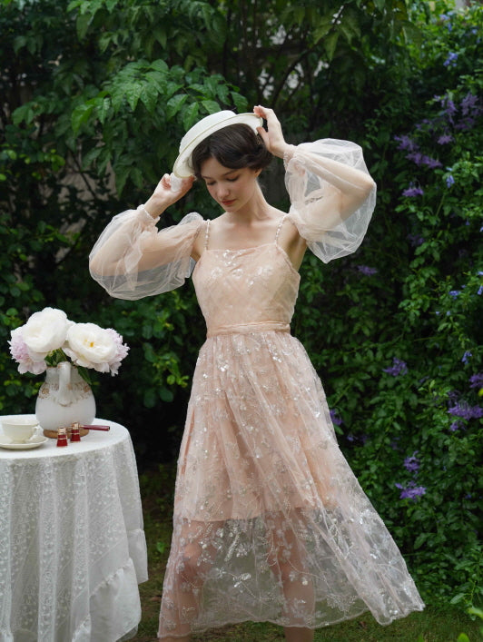 Model adjusts her hat while wearing a sparkly blush tulle dress with sheer puff sleeves, evoking whimsical fairycore elegance in a floral setting.