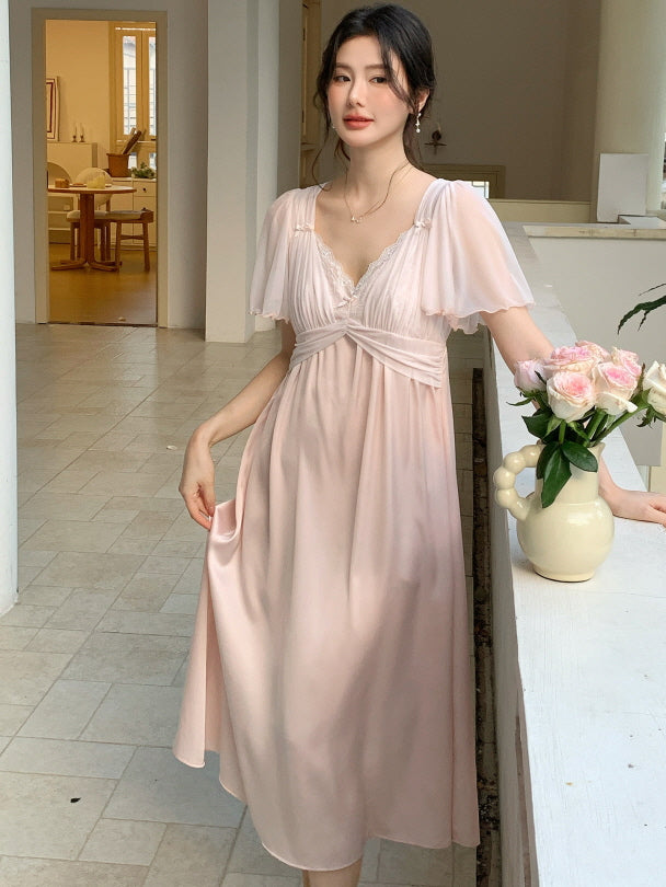 A woman stands beside a vase of roses in a pastel pink princesscore night dress with lace accents and a soft, flowing silhouette.