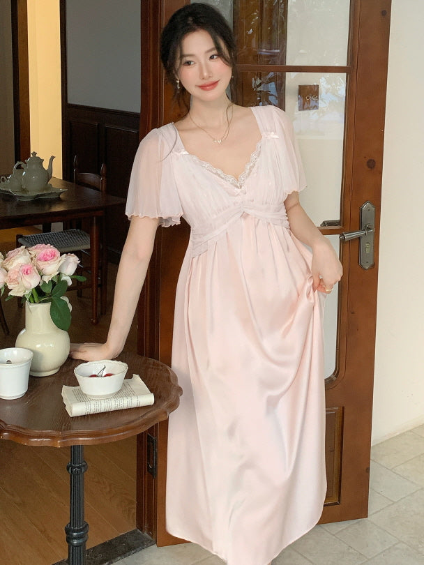 Leaning gently by a wooden door, the model wears a soft pink princess-style pajama dress with flutter sleeves, adding romantic charm to a vintage tea setting.