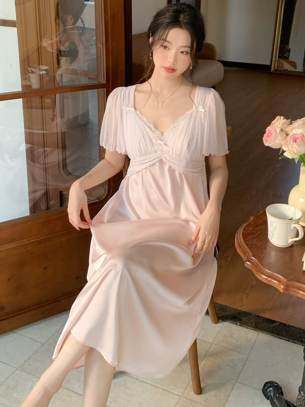 A woman in a dreamy princesscore loungewear dress sits by a wooden table, softly holding the hem of her pastel pink gown in a quiet morning scene.