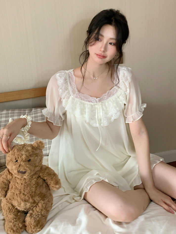 Model sitting on a bed with a teddy bear, wearing an ivory nightgown with lace neckline and soft chiffon layers for a romantic fairytale mood.