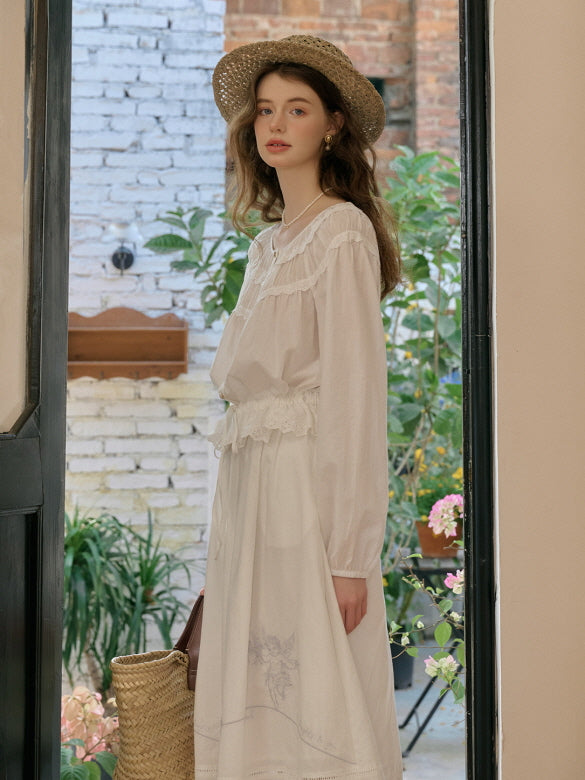 Woman wearing a vintage-inspired white blouse and skirt set with lace details, styled with a straw hat and a woven bag, standing in a garden doorway. Cottagecore outfit aesthetic.