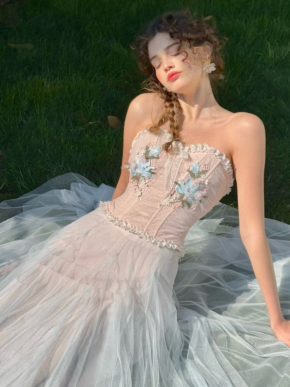 Woman in a pastel corset dress with blue floral embroidery and tulle skirt, reclining gracefully on grass in sunlight – dreamy romantic fairytale fashion