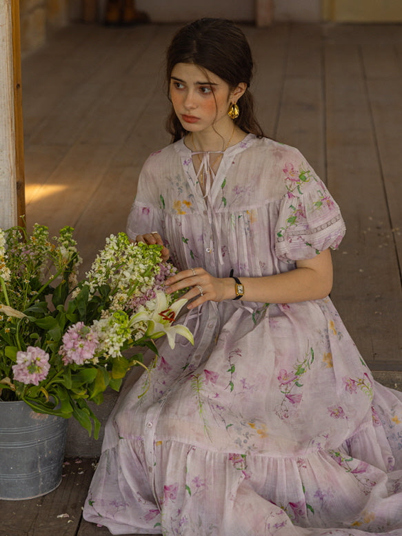 A model sitting on a wooden floor surrounded by fresh flowers, wearing a dreamy lavender floral dress with puff sleeves and a soft, ethereal design. The dress is adorned with delicate floral prints and a gathered waist for a romantic cottagecore aesthetic.