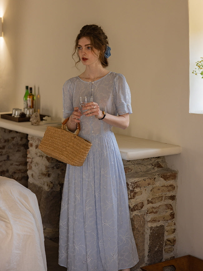 A woman holding a glass of water while wearing a blue eyelet lace dress with puff sleeves, styled with a woven basket bag. Cottagecore aesthetic in a rustic interior.