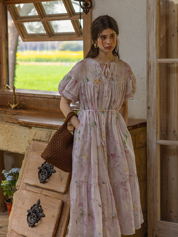 A model wearing a flowy, vintage-inspired floral dress in soft lavender tones, standing by a rustic window with countryside scenery in the background. The dress features puff sleeves, a delicate tie neckline, and a relaxed, tiered silhouette.