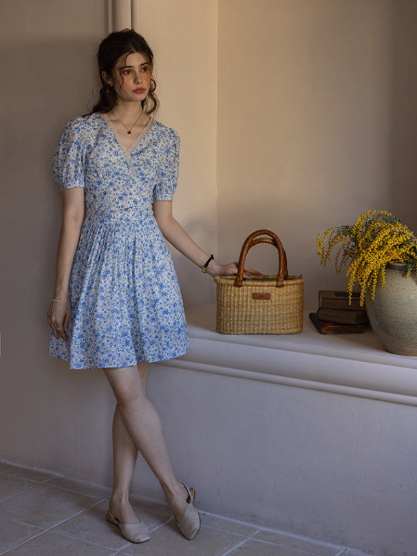 A woman standing against a neutral wall wearing a blue floral print short dress with puff sleeves, holding a woven basket. The scene evokes a vintage, cottagecore aesthetic with soft natural lighting.