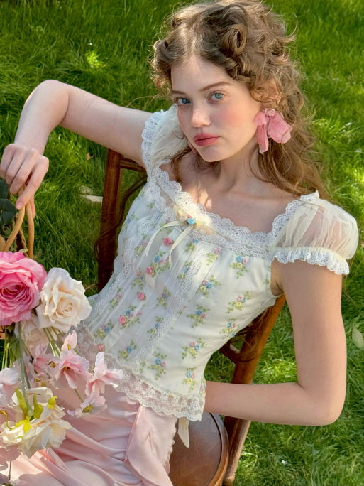 A model seated in a garden holding a basket of roses, wearing a floral lace corset top with sheer puff sleeves and pastel floral embroidery, styled with a pink ruched satin skirt for a dreamy, vintage-inspired look.