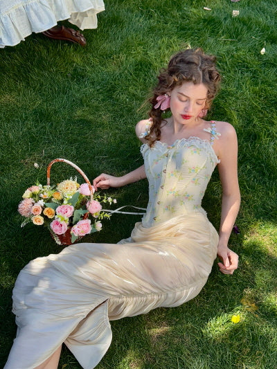 Cream ruched satin midi skirt styled with floral embroidered corset top, lying on the grass with a basket of pastel flowers – romantic fairytale garden outfit for women
