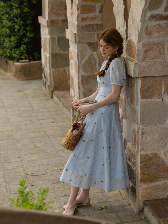 A woman leaning against a stone archway, dressed in a soft blue midi dress adorned with small green floral accents and a subtle check design. 