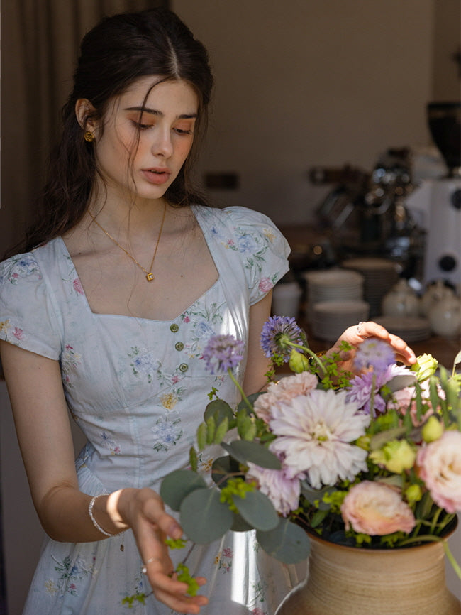 Charming floral dress with a fitted bodice, cap sleeves, and romantic flower prints, styled in a warm, vintage-inspired setting while arranging a bouquet of delicate blossoms.