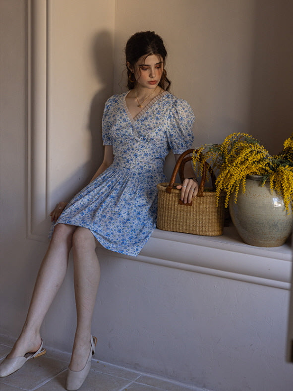 A woman seated against a neutral wall wearing a blue floral print short dress with puff sleeves, holding a woven basket placed beside her. The surrounding decor includes a large ceramic vase filled with bright yellow flowers, creating a warm, vintage-inspired ambiance.