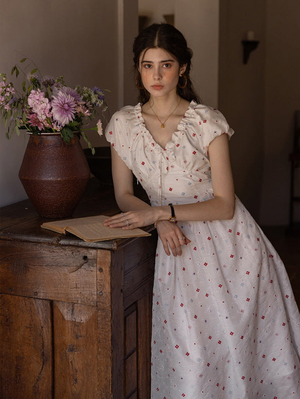 Woman in a cream floral print dress with ruffled neckline and delicate button detailing, leaning against a wooden cabinet with an open book and a vase of flowers, evoking a nostalgic, vintage atmosphere.