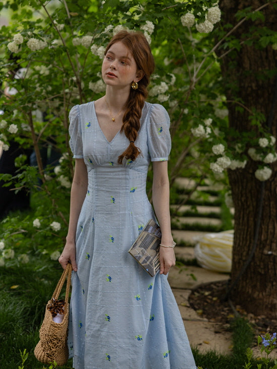 Woman standing amidst blooming white flowers, wearing a light blue floral midi dress with puff sleeves and a woven bag. The serene garden setting creates a romantic, cottagecore aesthetic.