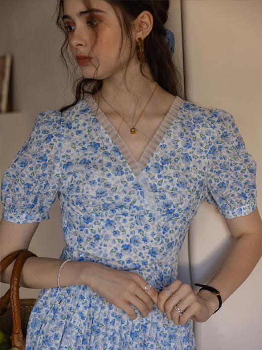 A woman wearing a blue floral dress with a V-neck lace trim, accessorized with gold jewelry and holding a woven basket, evoking a charming, vintage-inspired look.