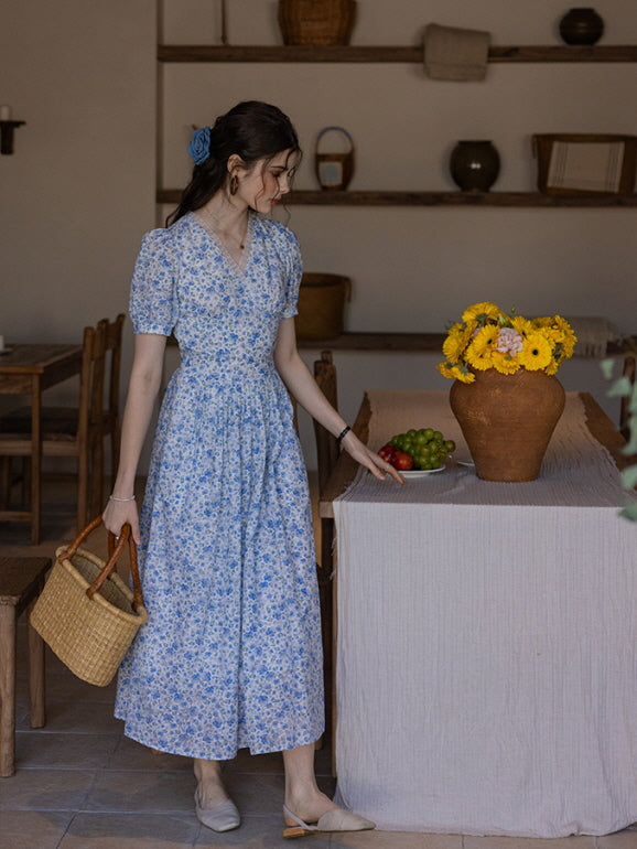 A woman wearing a blue floral print long dress, holding a woven basket while pointing at a table with a vase of yellow sunflowers. The vintage-inspired interior creates a nostalgic atmosphere.