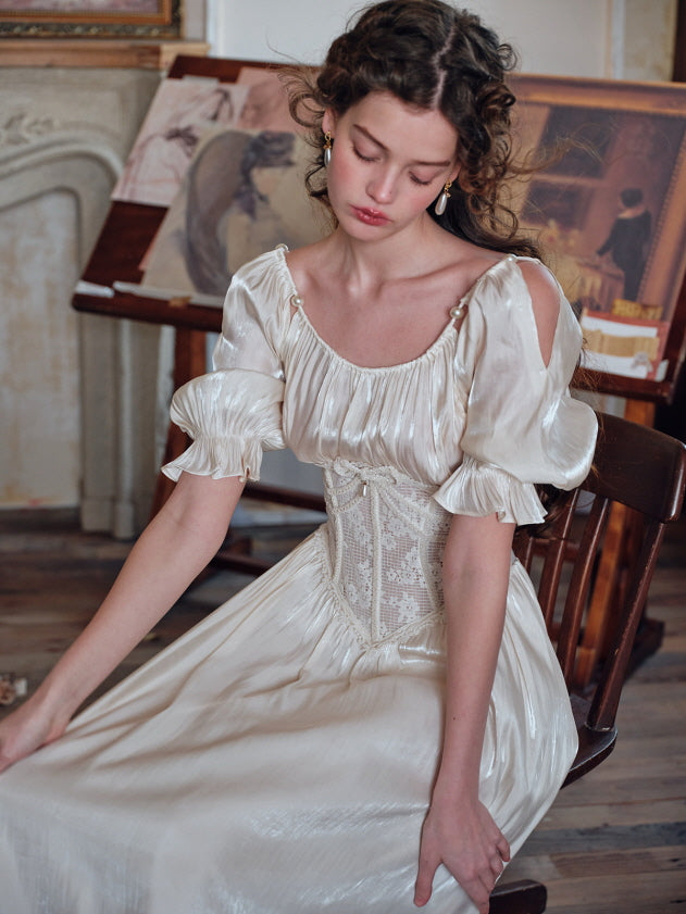 A woman sitting quietly in an ivory satin dress with delicate puff sleeves, shoulder cutouts, and a romantic lace corset, surrounded by classical paintings and antique decor.