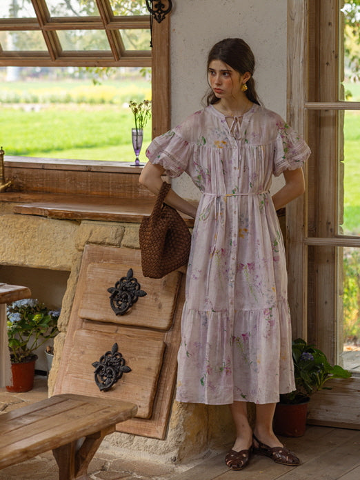 Model standing by a rustic kitchen window, wearing a light lavender floral tiered dress with puff sleeves and a drawstring neckline. The scene exudes a dreamy cottagecore aesthetic with a serene countryside view in the background.