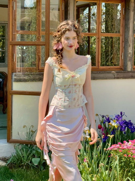 Romantic pastel pink ruched satin skirt with side ribbon detail, paired with a floral off-shoulder corset top, worn in a spring garden full of blooming flowers – fairytale vintage women’s fashion