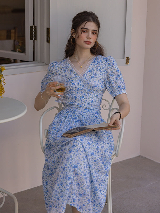 A woman seated on a white patio chair, wearing a blue floral print long dress, holding a glass of drink while reading a magazine against a soft pink wall, evoking a serene summer afternoon.