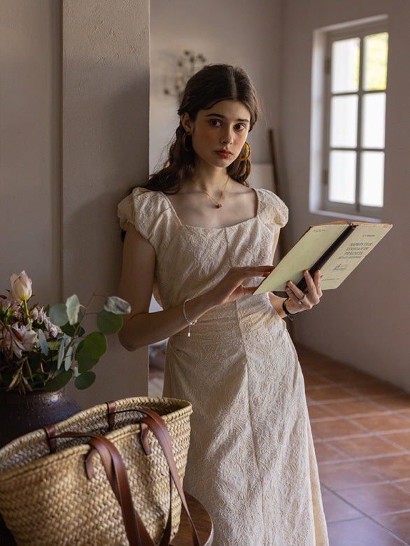 Woman in a cream-colored textured midi dress with a square neckline and puff sleeves, reading a vintage book while leaning against a wall in a softly lit, rustic room. A woven basket sits nearby, enhancing the cottagecore aesthetic.