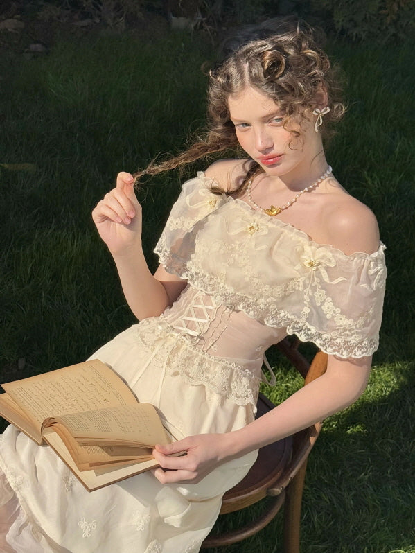 Woman in ivory lace off-shoulder corset dress reading a vintage book in a sunlit garden – romantic fairytale fashion with floral embroidery and ruffle details