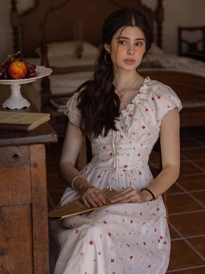 Young woman seated in a cream floral print dress with a ruffled neckline and puff sleeves, holding a vintage book while gazing thoughtfully to the side. The warm, rustic room enhances the nostalgic, cottagecore vibe.