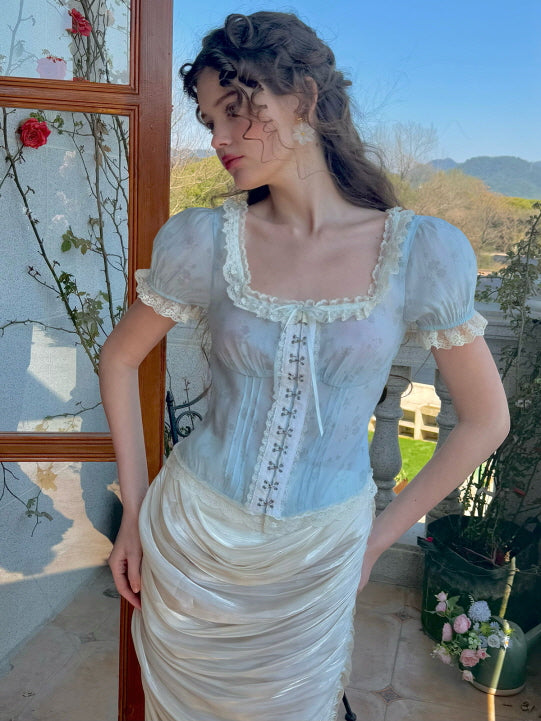 A model poses in a pastel blue floral corset top with lace trim and puff sleeves, styled with a ruched satin skirt, evoking a soft, vintage-inspired aesthetic against a garden backdrop with red roses.
