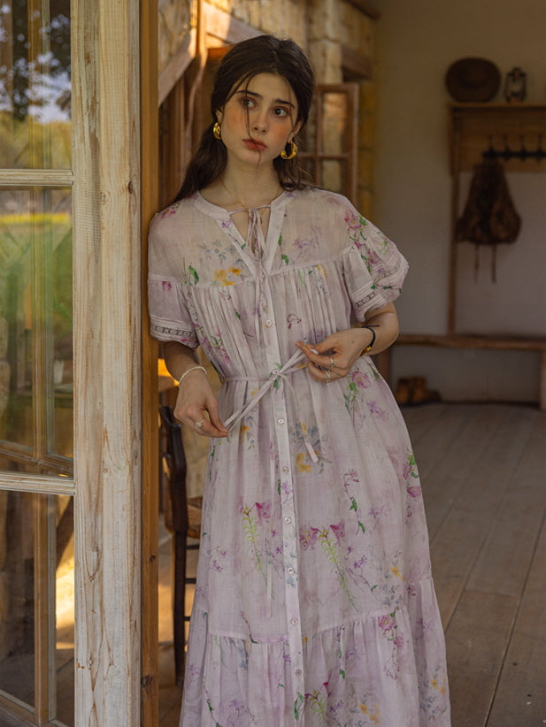 Model standing near a rustic wooden door in a cottage-inspired setting, wearing a light lavender floral print tiered dress with puff sleeves and a delicate tie neckline. The dress flows elegantly with a soft, romantic aesthetic.