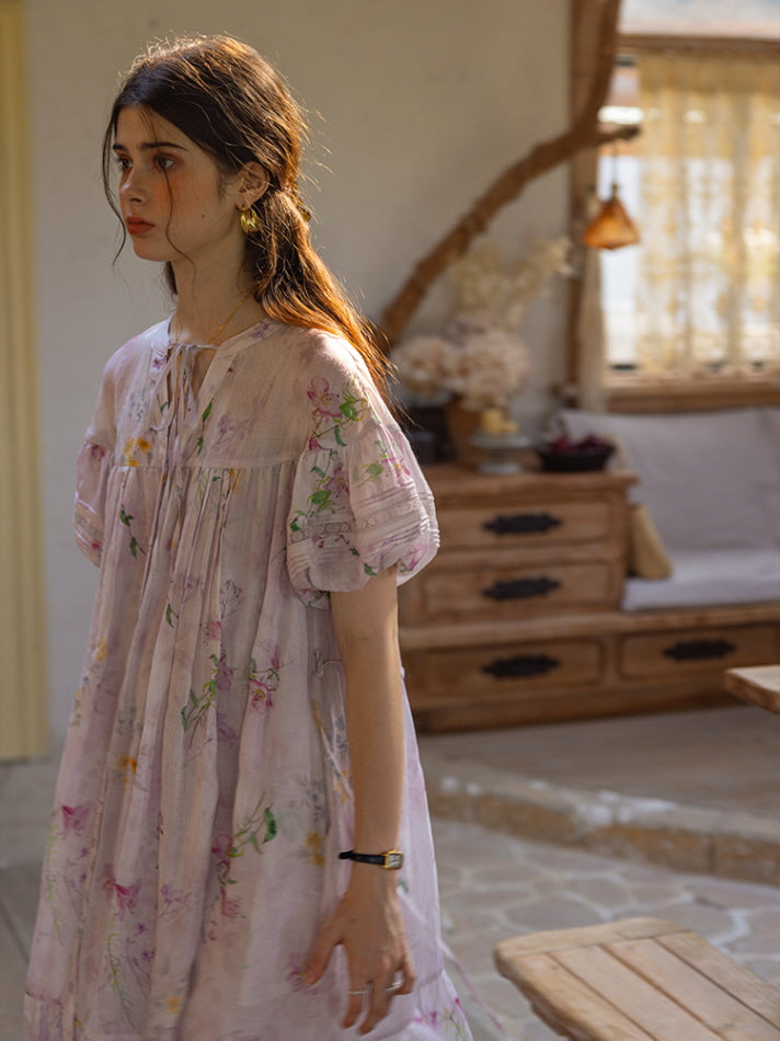 Model walking through a cozy, vintage-style room in a light lavender floral print dress with puff sleeves and a flowy, tiered silhouette. The dress exudes a dreamy, fairytale-inspired cottagecore vibe.