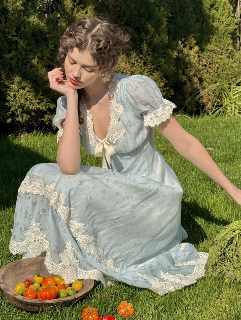 A woman sitting on green grass in a light blue lace-trimmed dress with puff sleeves and a bow, surrounded by cherry tomatoes in a wooden bowl, evoking a dreamy, countryside fairytale mood.