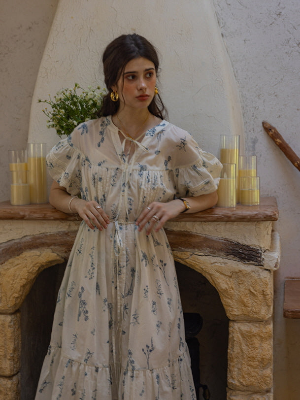 Woman standing in front of a stone fireplace wearing a cream floral print dress with blue botanical motifs and puff sleeves, surrounded by candles and rustic decor. The setting evokes a nostalgic, vintage-inspired ambiance.