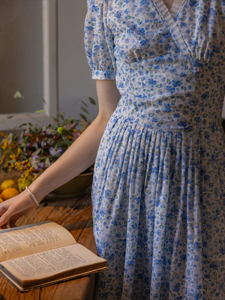 A close-up of a blue floral print dress with delicate pleats, styled with a vintage book on a wooden table, creating a cozy cottagecore ambiance.