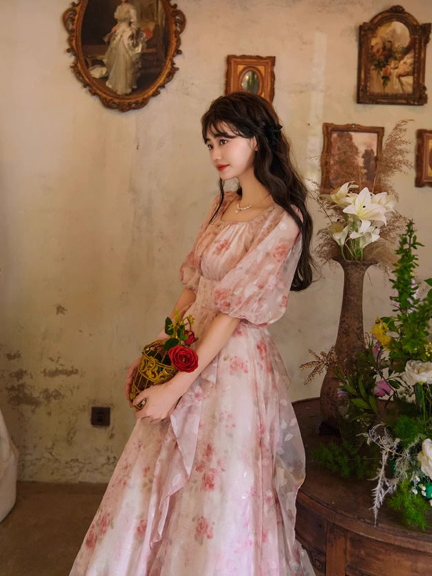 A fairytale-inspired gown with soft pink florals, a square neckline, and flowing silhouette, creating a princesscore look against a vintage floral backdrop.