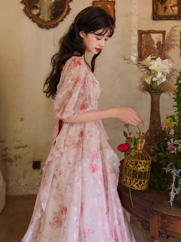 A delicate floral gown with soft pink blooms, cascading ruffles, and a romantic square neckline, paired with a gold birdcage for a fairytale touch.
