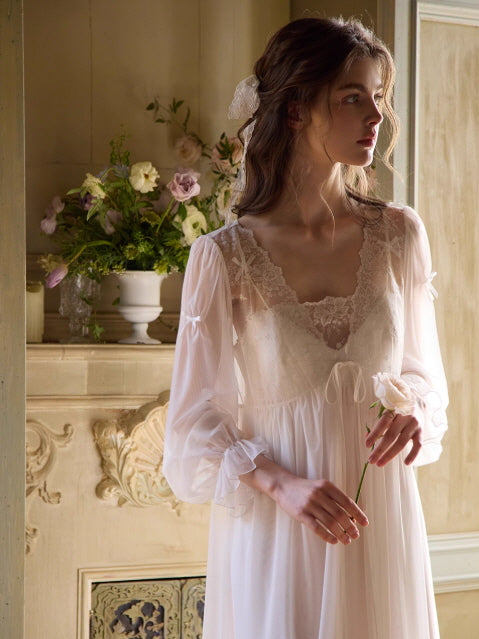 A woman in a sheer white lace-trimmed gown stands by a vintage mantelpiece, holding a pale flower in a soft fairytale-inspired scene.