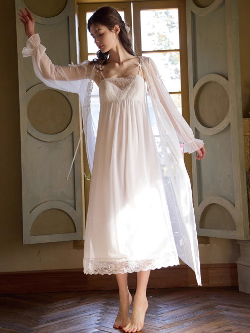 Wearing a flowing white nightgown with lace hem, the model lifts her robe slightly as sunlight streams through vintage shutters in a dreamy moment.