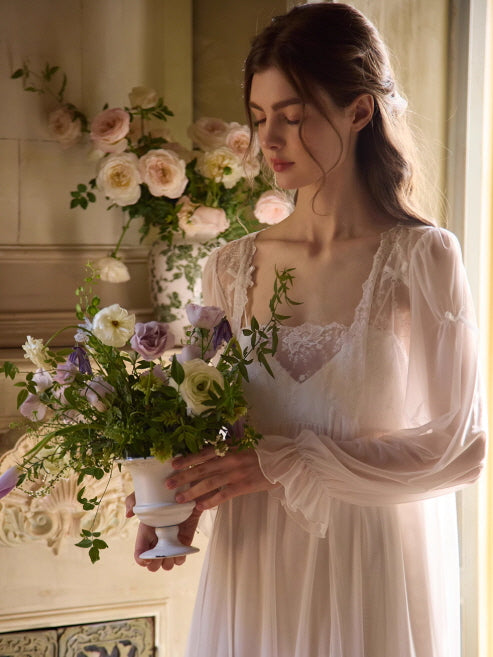 A woman in a sheer vintage-inspired gown gently holds a white vase of garden flowers, bathed in soft morning light with a fairytale touch.