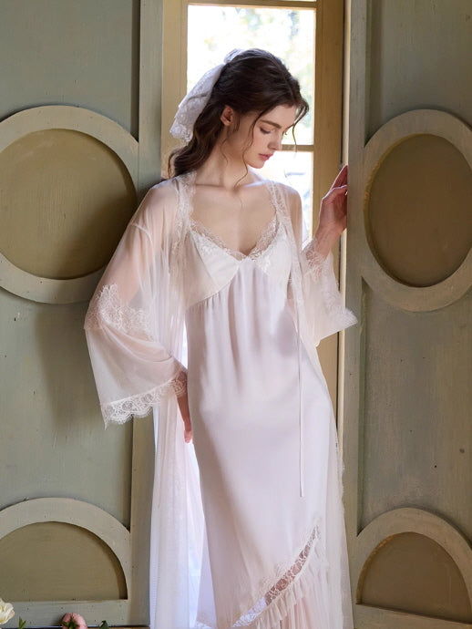 Wearing a princesscore-inspired white loungewear set, the model stands beside a sunlit window in a sheer lace robe over a delicate slip dress.