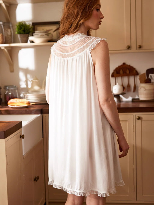 A woman wears a flowy white lace-trimmed nightgown with a sheer yoke and ruffled hem, styled in a soft vintage-inspired cottage kitchen.