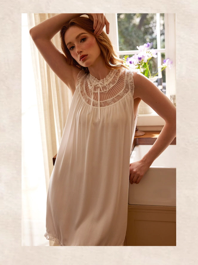 A young woman poses gracefully in a white lace-trimmed nightgown by the window, evoking quiet vintage elegance in a warm cottage setting.