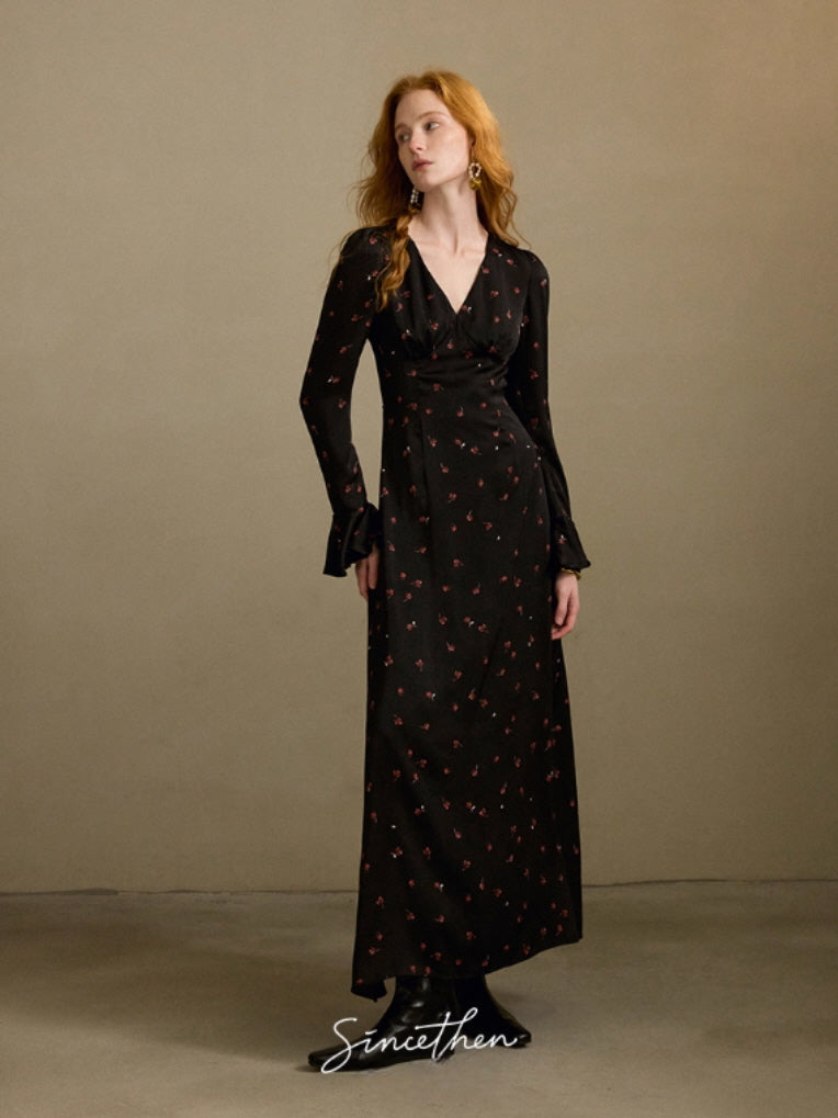 Model wearing a long black floral maxi dress with a vintage V-neck silhouette and subtle red blossom pattern, standing in a neutral studio setting.