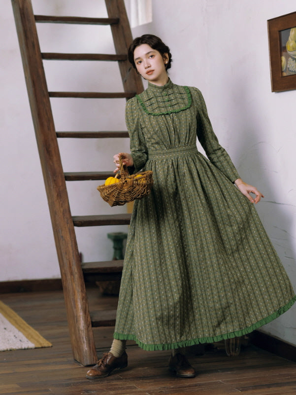 Model wearing a long green vintage prairie dress with a ruffled bib detail, standing indoors with a woven basket in hand for a classic cottagecore aesthetic.