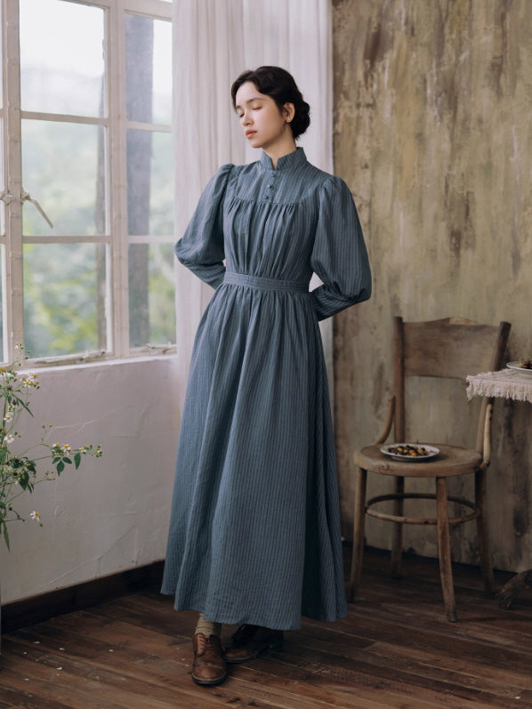 Model in a blue cottagecore high-neck pleated long dress standing by a window, showcasing romantic vintage aesthetic fashion.