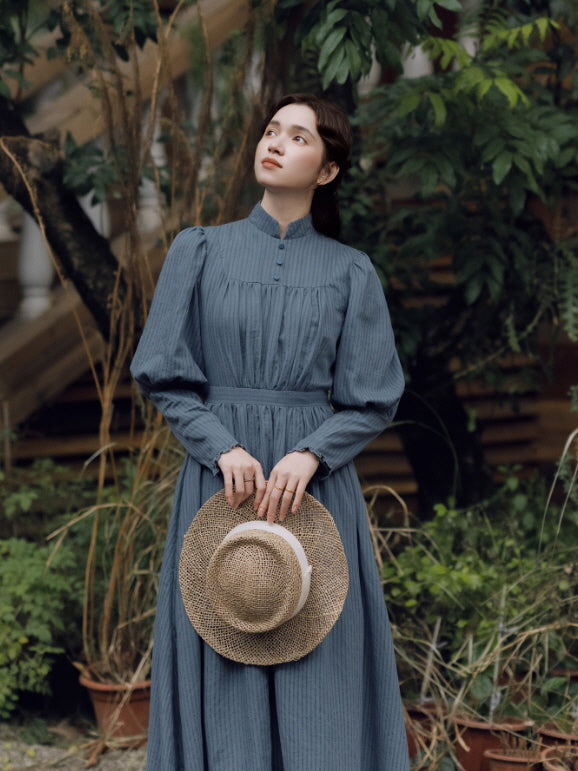 Woman wearing a vintage-inspired blue long-sleeve maxi dress with pintuck details and a high collar, holding a straw hat while standing in a garden.