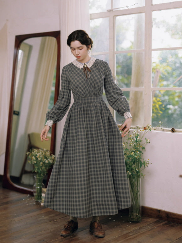 Model wearing a grey check vintage long dress with lace collar and ribbon brooch, standing by a bright window in a cottage-style room – cottagecore and fairytale aesthetic outfit.
