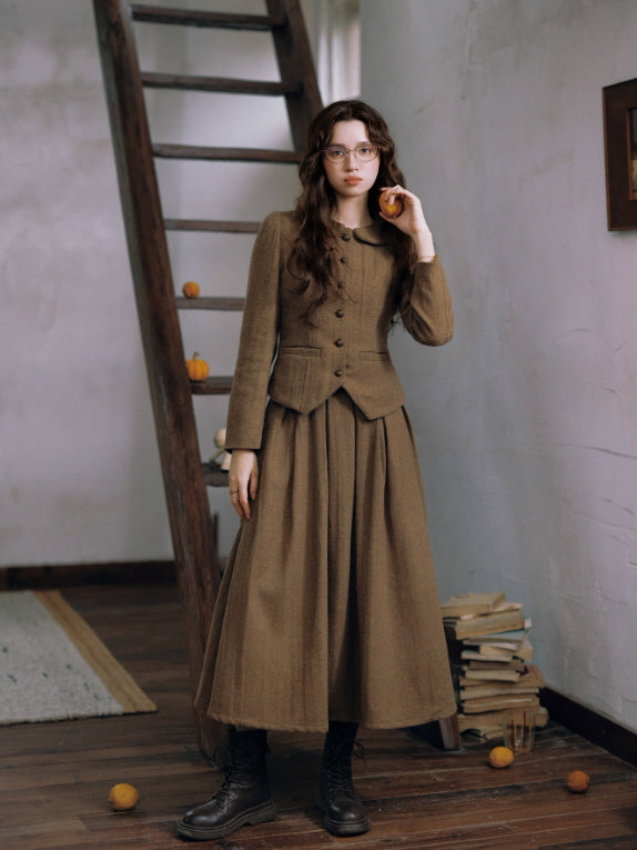 woman in a vintage brown tailored jacket and long pleated skirt, standing indoors beside a wooden ladder and scattered small pumpkins, styled with round glasses for a nostalgic cottagecore aesthetic