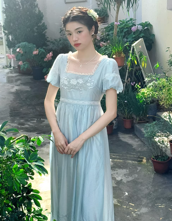 Close-up of a woman in a light blue cottagecore dress featuring white rose embroidery and delicate lace trim on a square neckline.