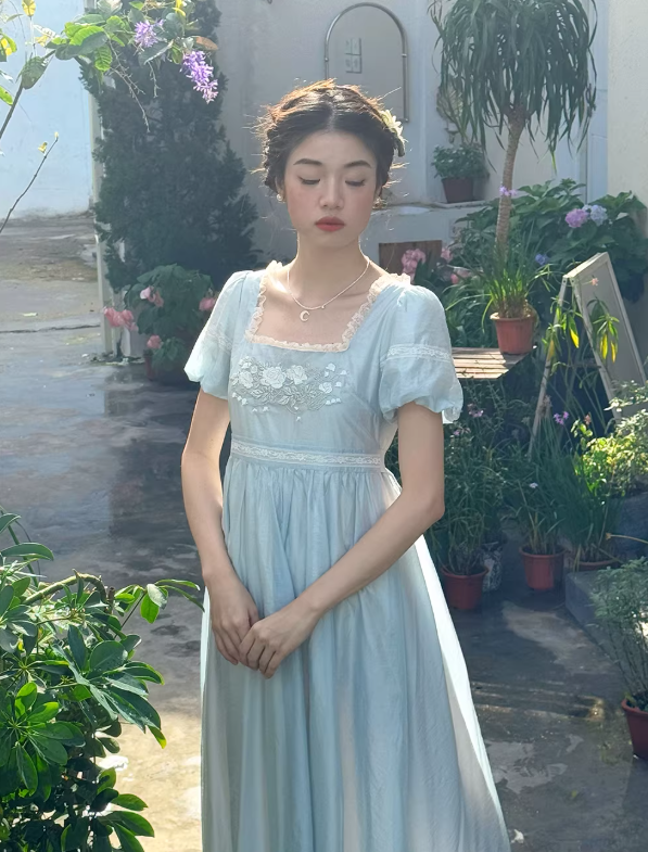 Ethereal woman with closed eyes wearing a fairytale-inspired pale blue gown with puff sleeves in a lush, sunlit garden setting.