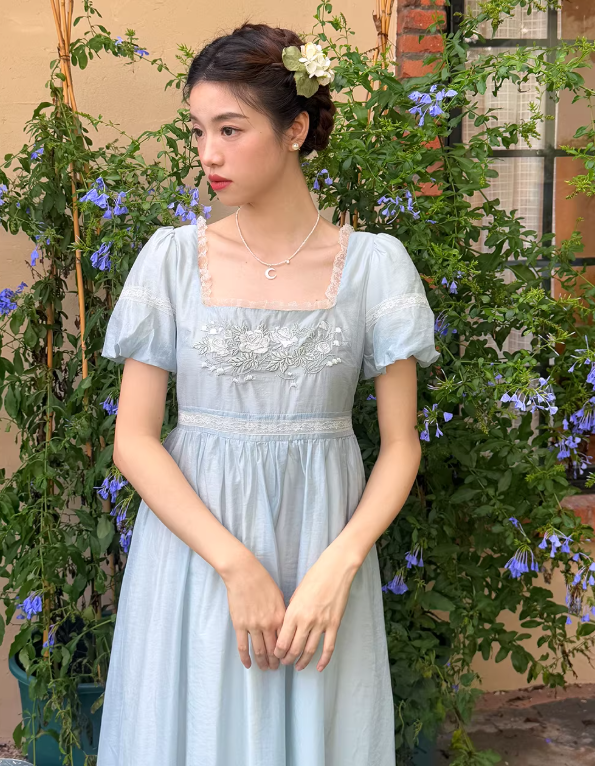 Detail view of a light blue cottagecore dress featuring intricate white rose embroidery, vintage lace trim on a square neckline, and a dainty moon necklace.